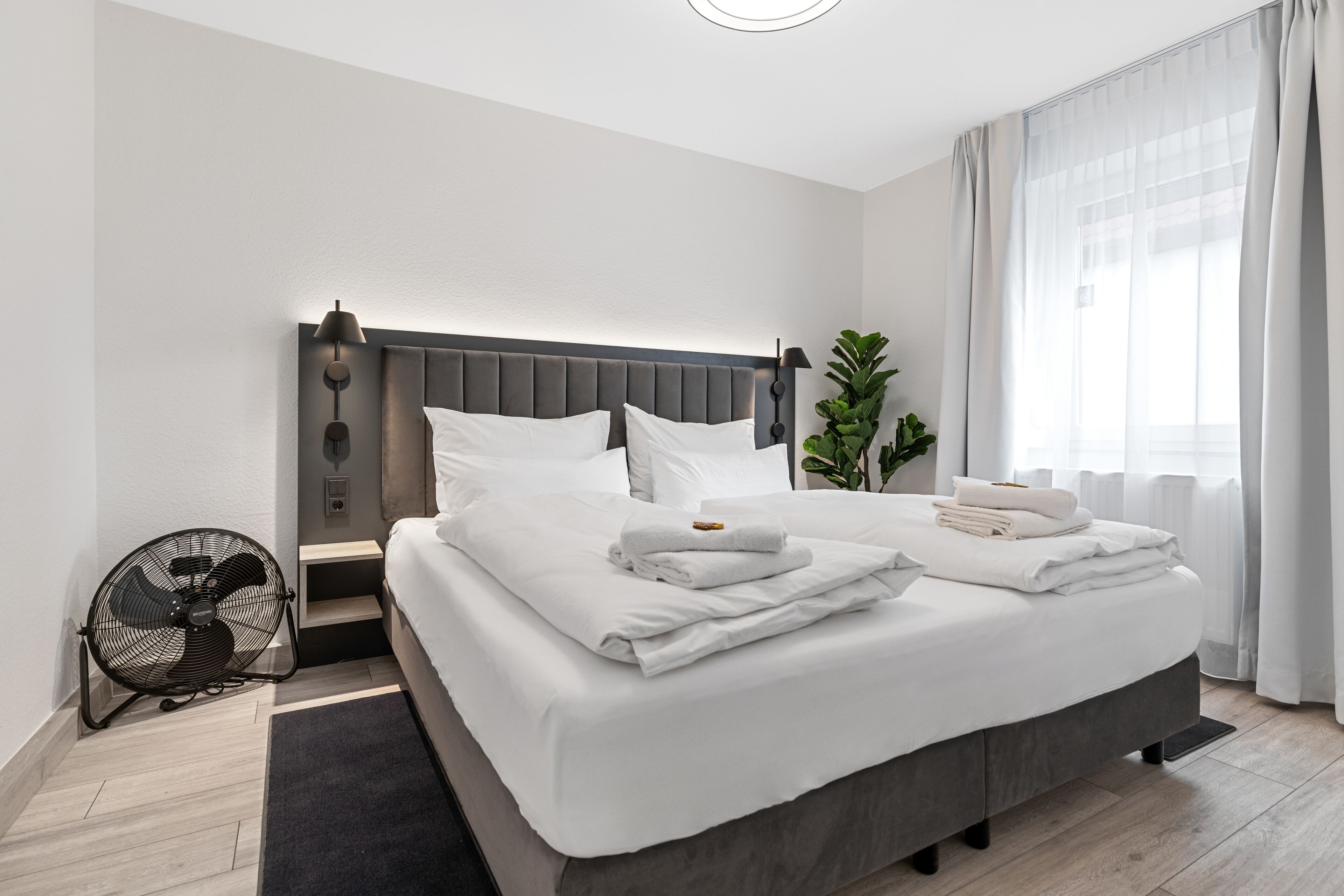 Ein modernes Hotelzimmer mit einem großen Bett, einem Nachttisch, einem Ventilator und einem Fenster mit Vorhängen.