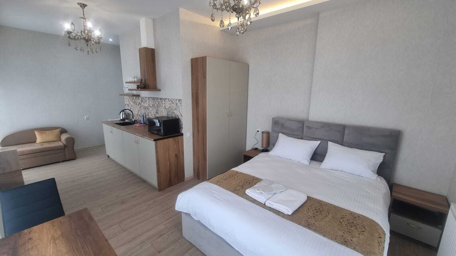 A modern hotel room with a large bed, a separate seating area, a kitchenette, and a chandelier.