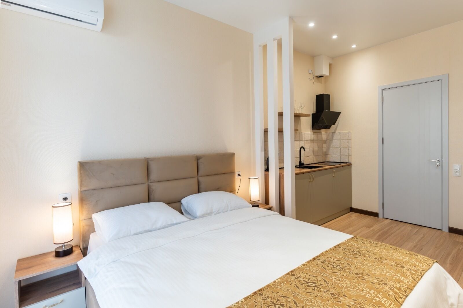 A hotel room with a bed, bedside lamps, a small bedside table, a kitchen area with a sink and cabinets, and a door.