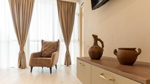 A room with a wooden floor, a beige armchair with a patterned cushion, a wooden cabinet with two drawers, and a wall-mounted flat-screen TV. - Sky By The River (Tbilisi)