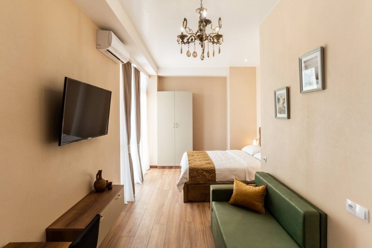 Ett modernt hotellrum med en platt-TV, en grön soffa, en säng med ett mönstrat överkast och en kristallkrona.