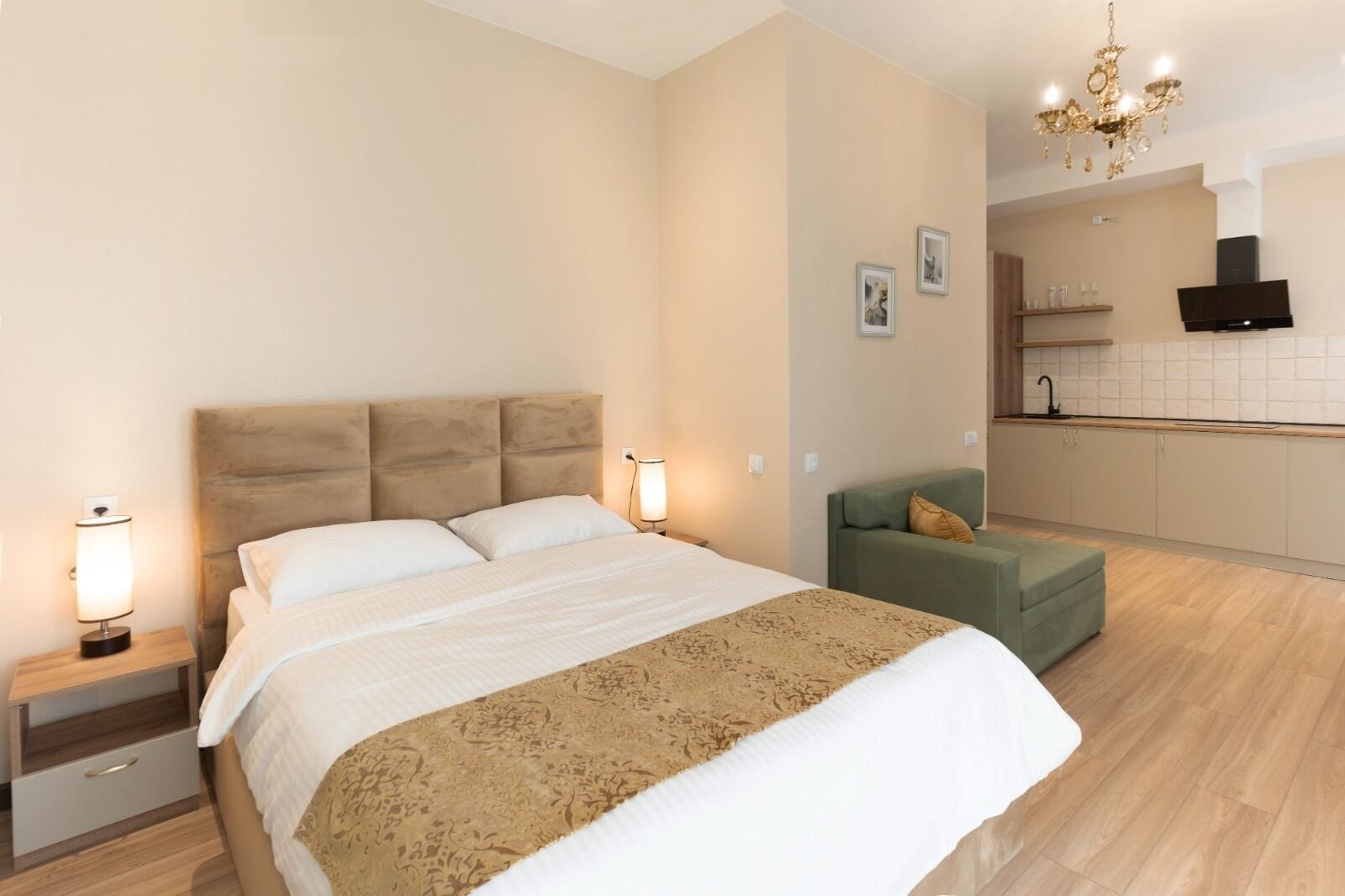 A bedroom with a large bed, bedside tables, a green armchair, a chandelier, and a kitchenette in the background.