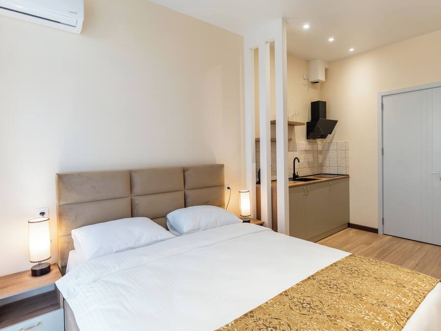 A bedroom with a bed, bedside tables, a nightstand, a lamp, a patterned throw, a wardrobe, and a kitchen area with a sink and cabinets.