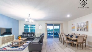 Ein geräumiges Wohnzimmer mit Sofa, Couchtisch und Fernseher. Es gibt einen Essbereich mit Tisch und Stühlen. Das Zimmer verfügt über ein großes Fenster und einen Blick ins Freie.