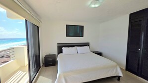 A bedroom with a bed, a nightstand, a window with a view of the beach, and a ceiling light. - Oceanfront white sand beach condo  (Cancun)