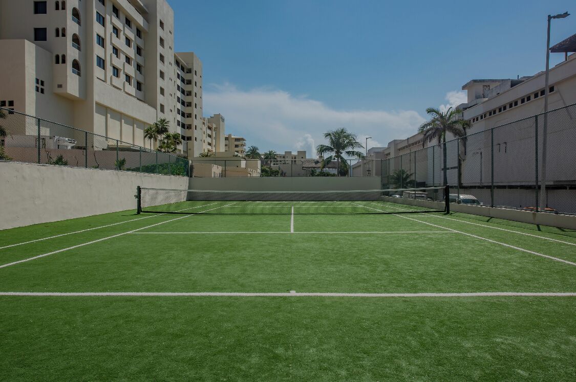 An outdoor tennis court with a net, surrounded by a fence, and adjacent to a building with multiple floors and balconies.