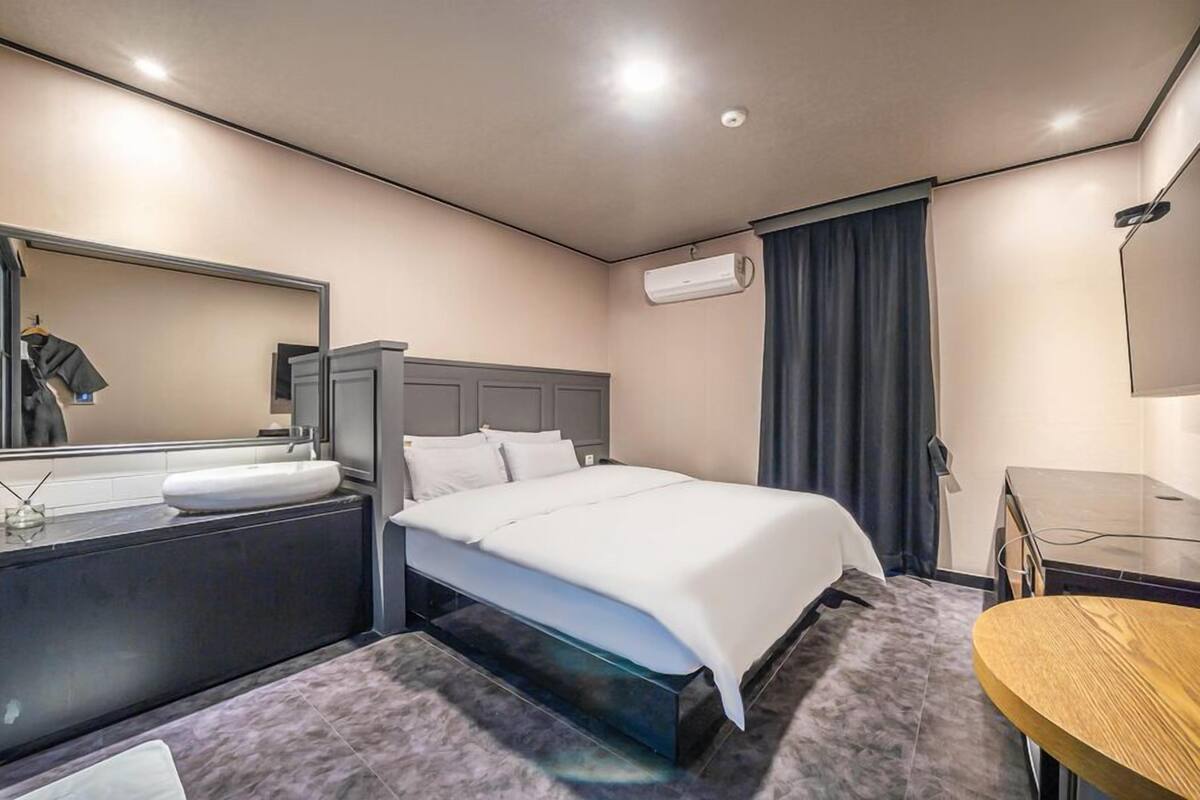 Habitación de hotel con cama, mesita de noche con lámpara, espejo, televisor y una pequeña mesa redonda.
