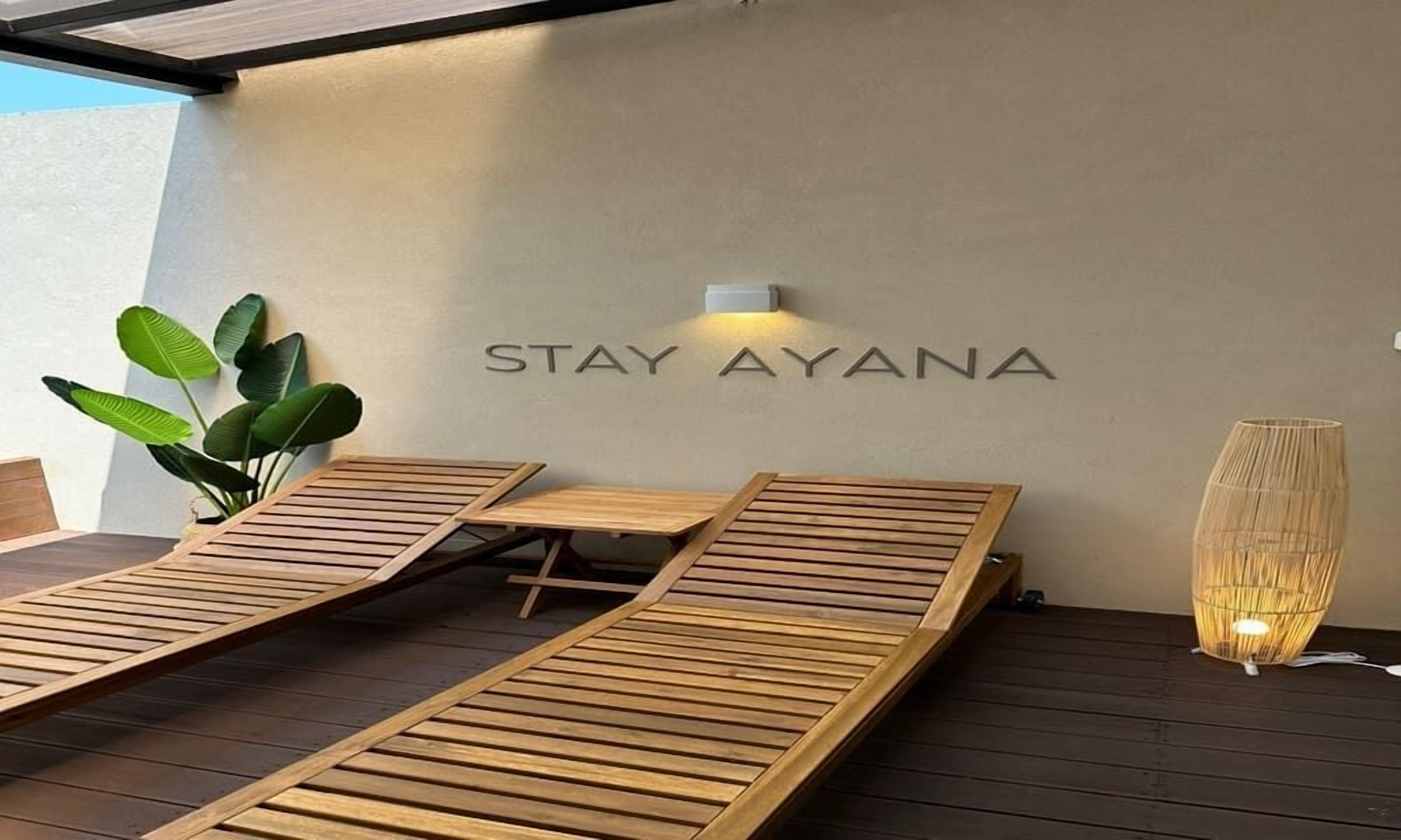 A modern outdoor seating area with wooden loungers and a wall-mounted sign that reads "STAY AYANA".