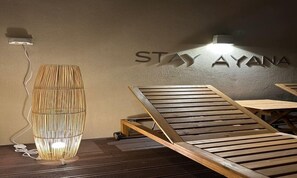Een kamer met een wandbord waarop 'STAY AYANA' staat, een houten lounge stoel en een cilindrische lamp met een geweven ontwerp.