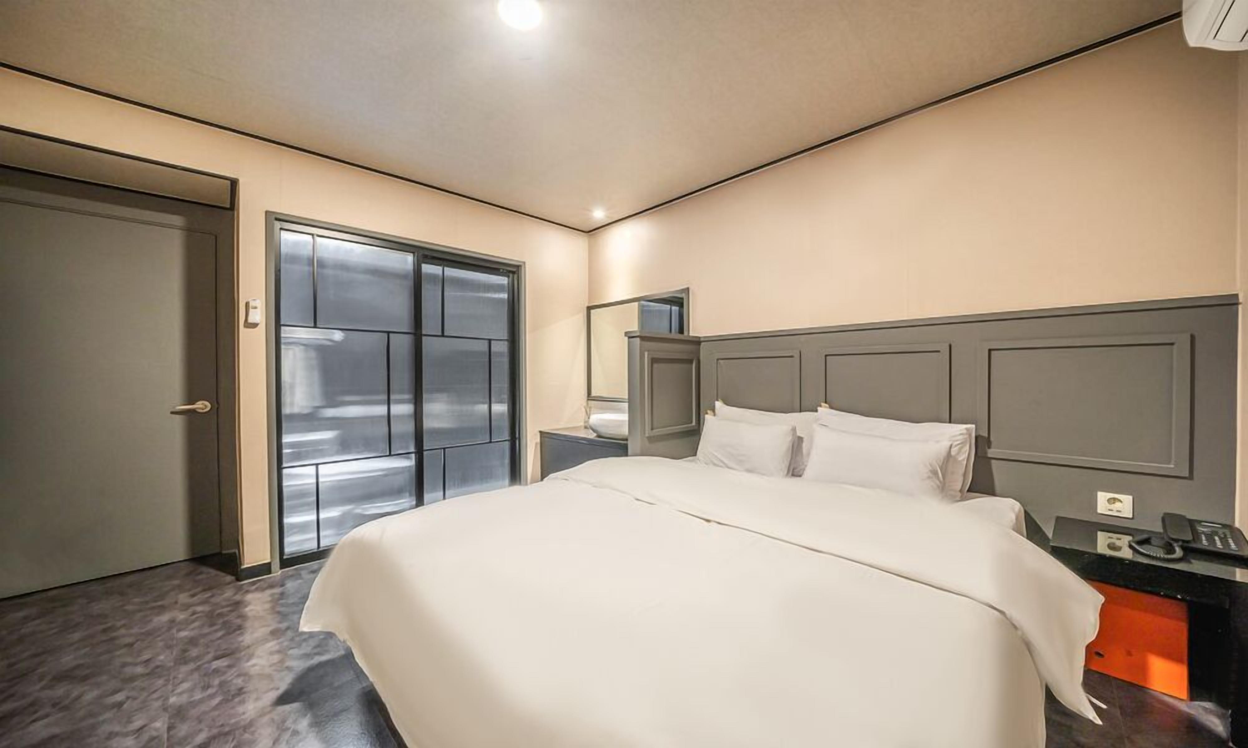 A modern hotel room with a large bed, a bedside table, a nightstand, a mirror, and a wardrobe.