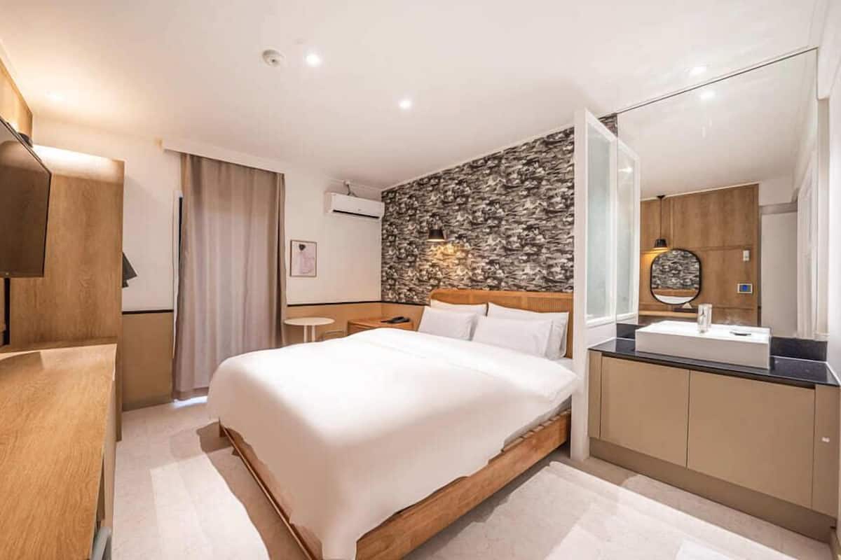 Una habitación de hotel moderna con una cama grande, un cabecero de madera, una pared decorativa con textura de piedra, una mesita de noche pequeña, un espejo y un baño con lavamanos y tocador.
