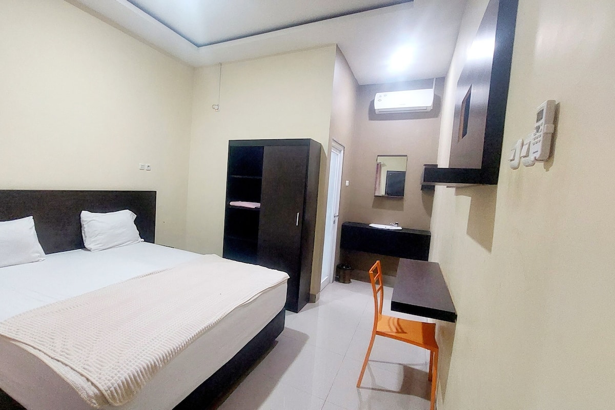A hotel room with a bed, a desk, a chair, a wardrobe, a mirror, and a small trash bin.