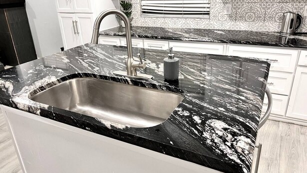 Una cocina con encimera de mármol negro, pileta de acero inoxidable y muebles blancos.