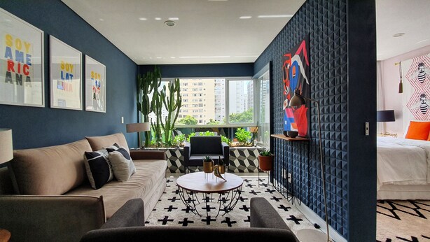 A modern living room with a sofa, coffee table, and chairs. There are framed pictures on the wall, a large window, and a view of buildings outside. - Moderno e Vibrante na Augusta (São Paulo)
