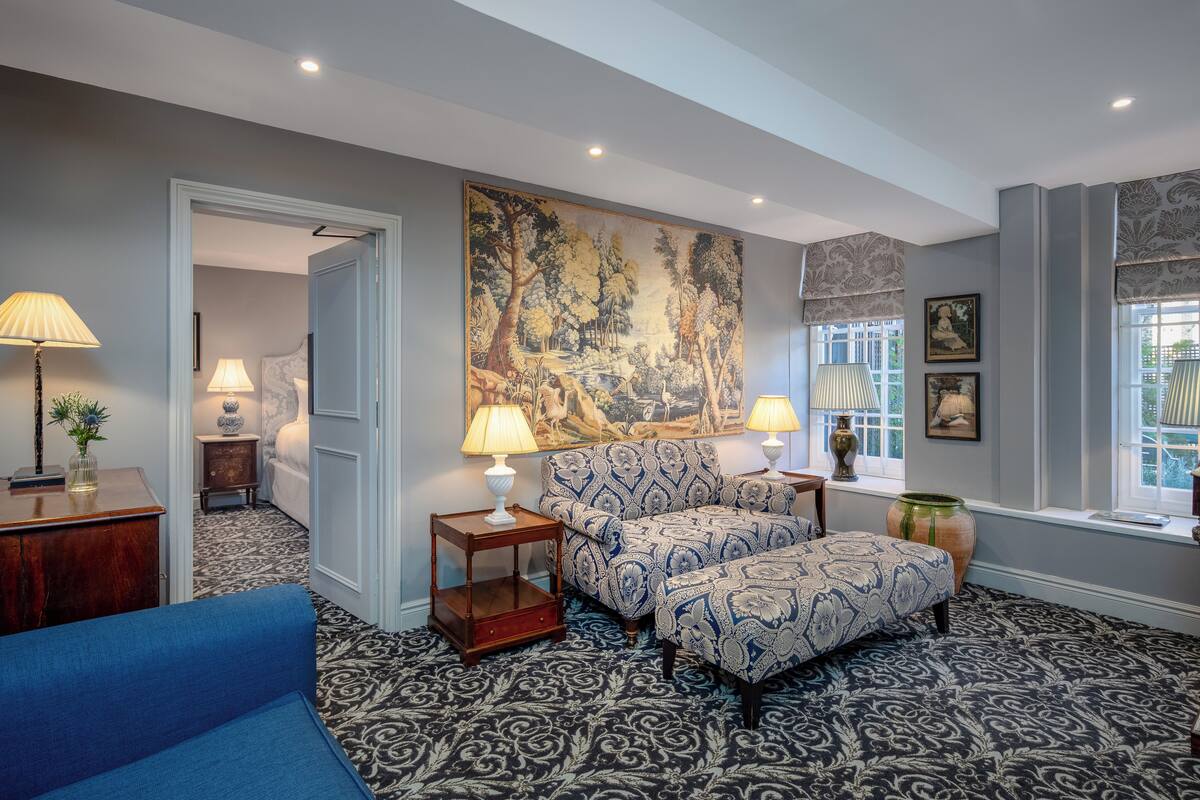 A room with a patterned carpet, a blue sofa, a patterned armchair, a side table with a lamp, a large painting on the wall, and a window with a view.