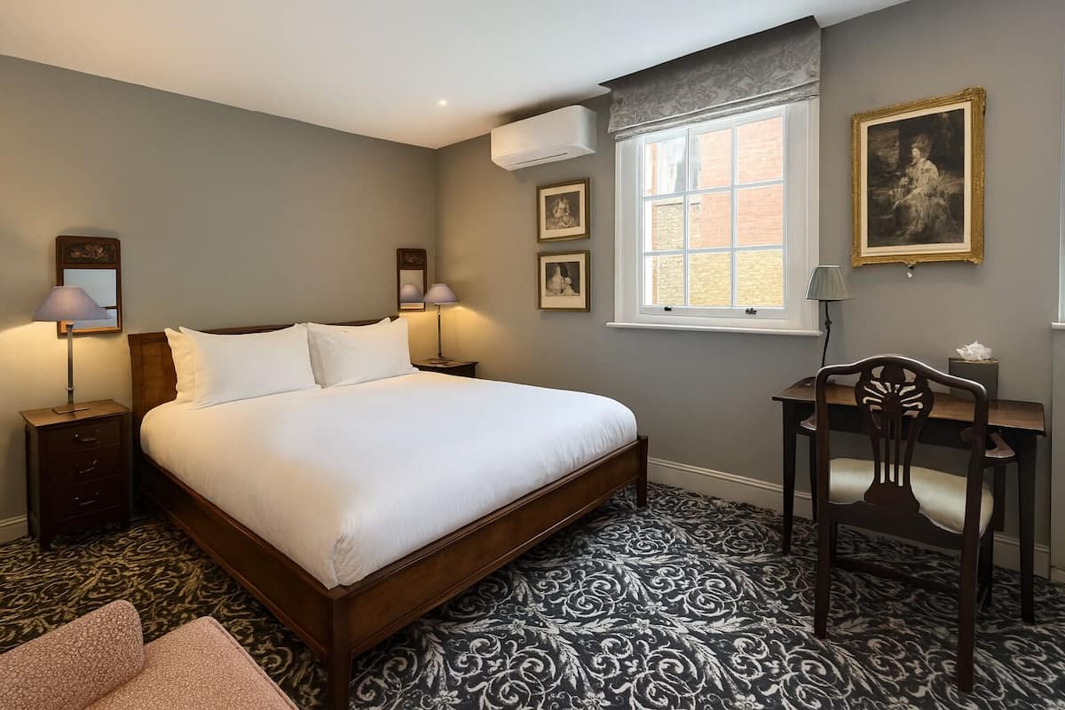 A hotel room with a large bed, bedside lamps, a desk with a chair, a window with a view, and artwork on the walls.