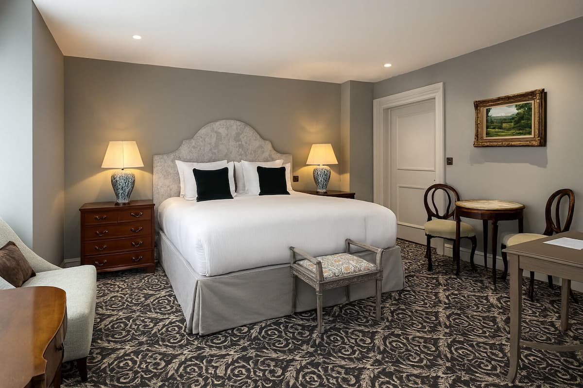 A hotel room with a large bed, two bedside tables with lamps, a chair, a small table, and a framed picture on the wall.