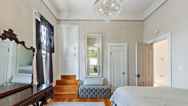 A bedroom with a large bed, a dresser, a mirror, a chandelier, and a staircase leading to another room. - Barclay Klum House by WanderLodges (Ashland)