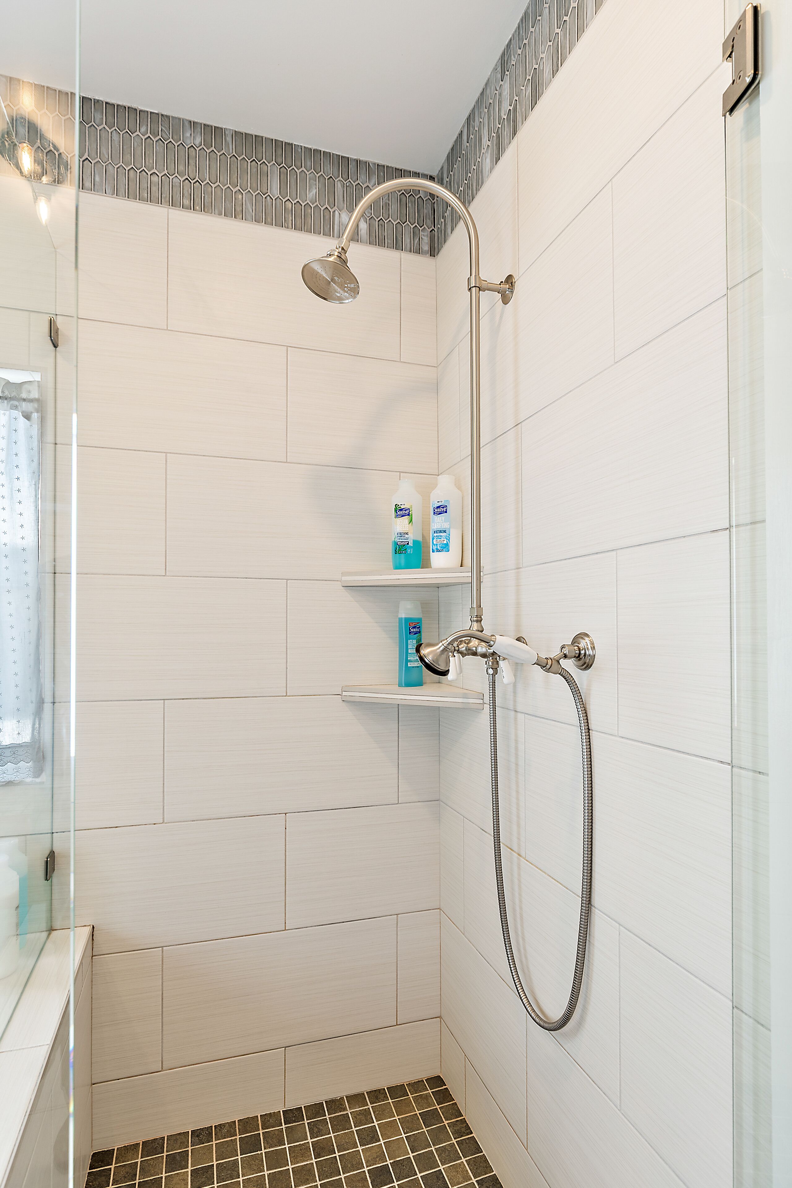 A shower with a handheld showerhead and a wall-mounted faucet, a glass shower enclosure, and a shelf with toiletries.