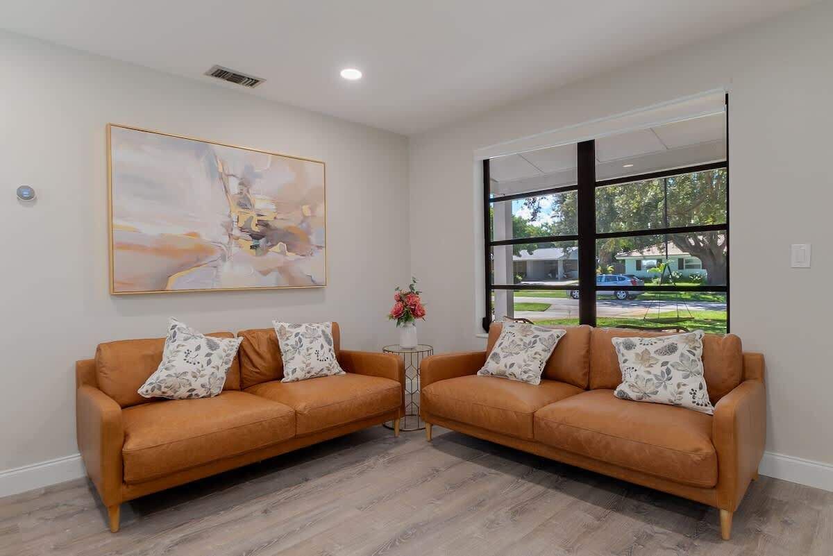 A room with two brown leather sofas, a small round table with a vase of flowers, a large abstract painting on the wall, and a large window with a view of the outside.