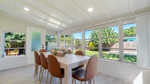 Un comedor con una mesa blanca y sillas marrones, ventanas amplias y vista a un jardín.