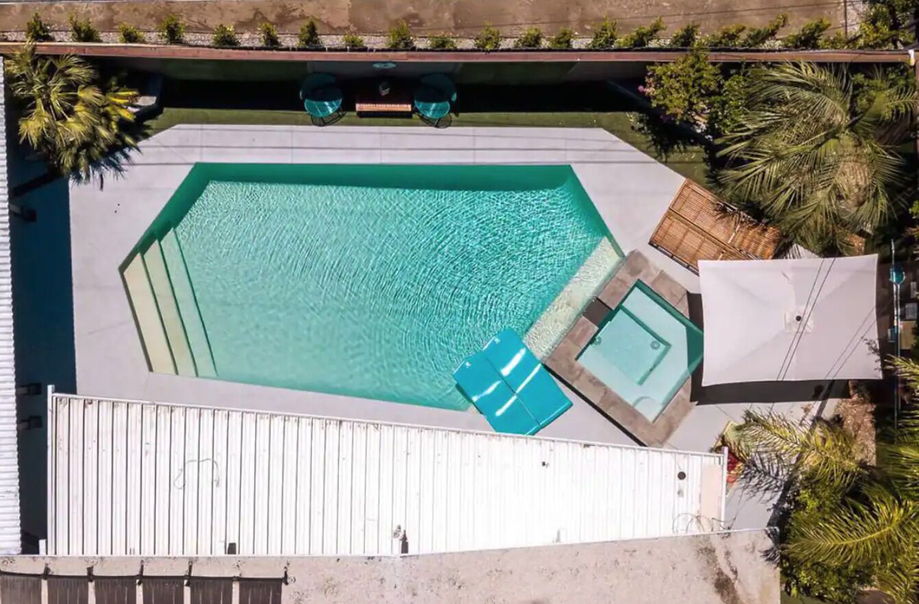Vista aérea de una piscina rectangular con un pequeño jacuzzi contiguo, rodeada por una valla blanca y palmeras.