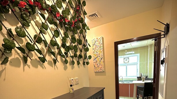 A hallway with a dark wooden cabinet, a wall-mounted plant with red flowers, and a tiled floor leading to a kitchen area. - Hello Hostel (Dubai)