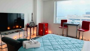 Une chambre d’hôtel avec un lit, une télévision, une petite table avec une chaise rouge, un meuble rouge et une fenêtre donnant sur la ville.