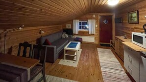 Um quarto de sótão acolhedor com teto de madeira, um sofá, uma pequena mesa, uma área de cozinha com micro-ondas e uma janela com cortinas.