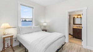 A bedroom with a bed, a nightstand with a lamp, a window with a view, and a door leading to a bathroom. - The Night Owl Perch (Stanwood)