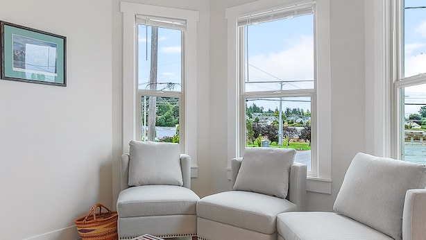A modern living room with two sofas, a coffee table, and a view of a residential area through large windows. - The Night Owl Perch (Stanwood)