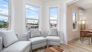 A modern living room with a sectional sofa, a wooden coffee table, and a dining area with a table and chairs. Large windows offer views of the outside. - The Night Owl Perch (Stanwood)