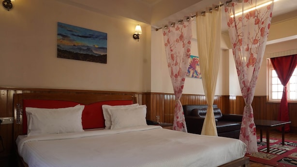 A neatly made bed with white linens, a wooden headboard, and red pillows. There is a painting on the wall, a lamp, and a window with red curtains. - Hotel Aster (Gangtok)