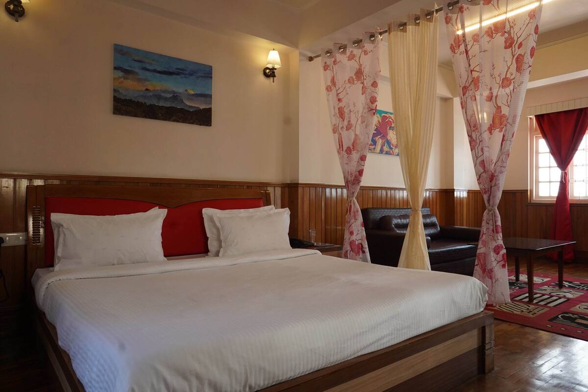 A neatly made bed with white linens, a wooden headboard, and red pillows. There is a painting on the wall, a lamp, and a window with red curtains.