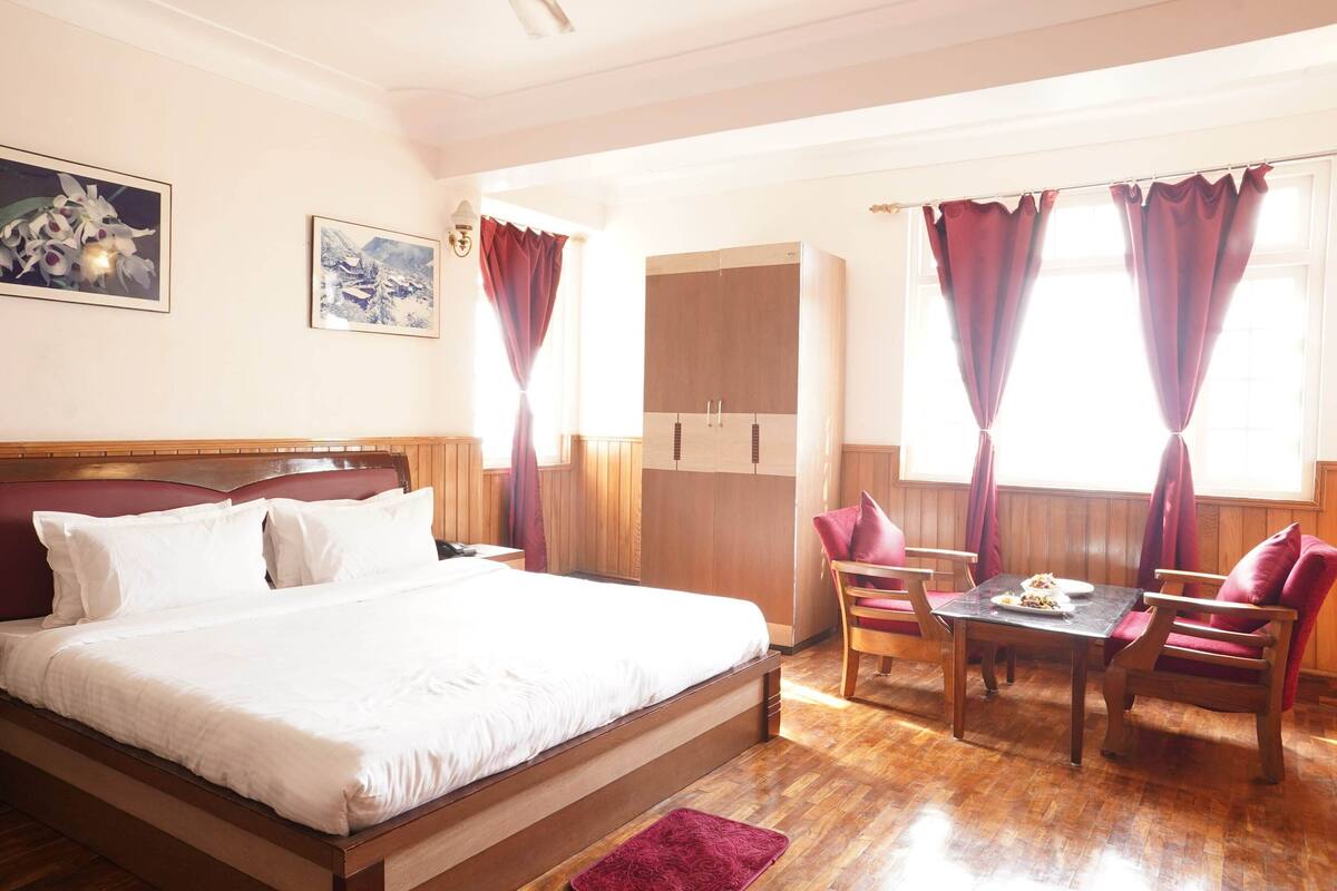 A hotel room with a large bed, a wooden table, two chairs, a small table with a plate, and a wardrobe.