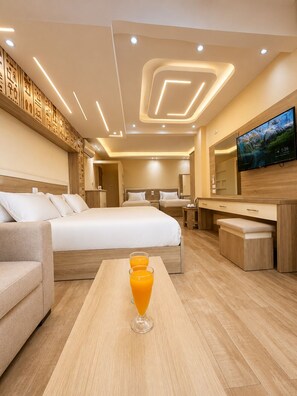 A modern hotel room with a large bed, a sofa, a wooden table with two glasses of orange juice, a TV, and a mirror.