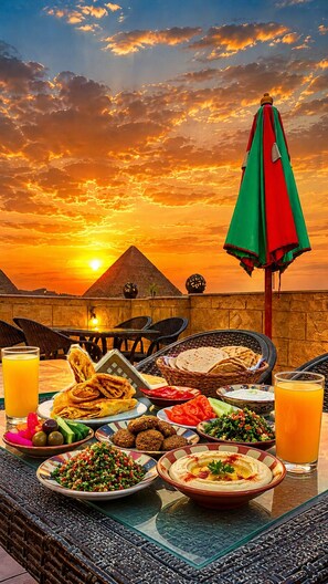A table set with various dishes and drinks, a wicker chair, and a view of pyramids at sunset.