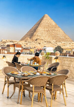 A dining area with wicker chairs and a table set for breakfast, overlooking a pyramid. - Hassan Pyramids Hotel (Giza)