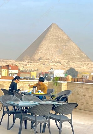 A dining area with a table and chairs, a pyramid in the background, and a stone wall. - Hassan Pyramids Hotel (Giza)