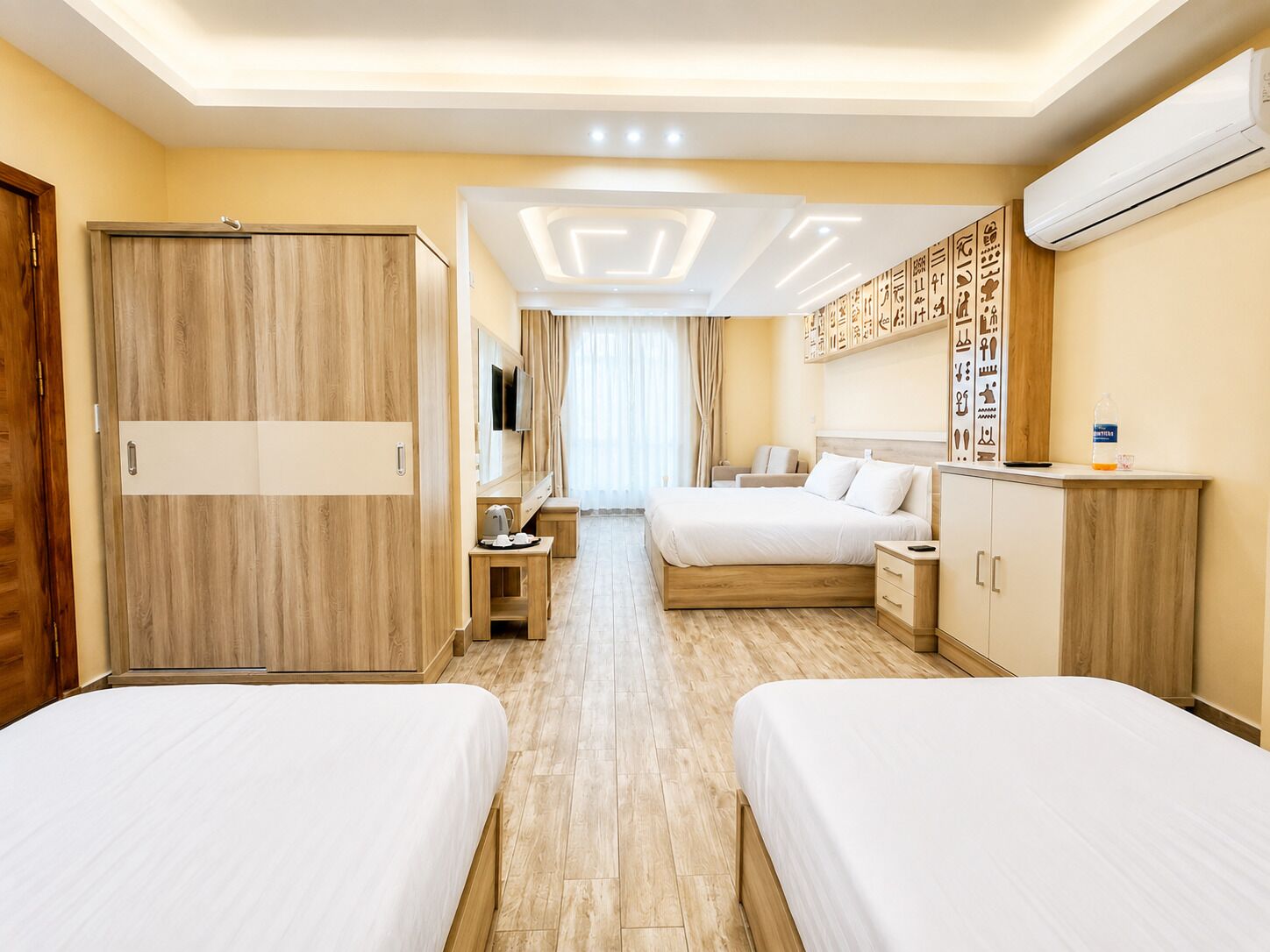 A hotel room with two beds, a desk, a TV, a minibar, and a wall with hieroglyphs.
