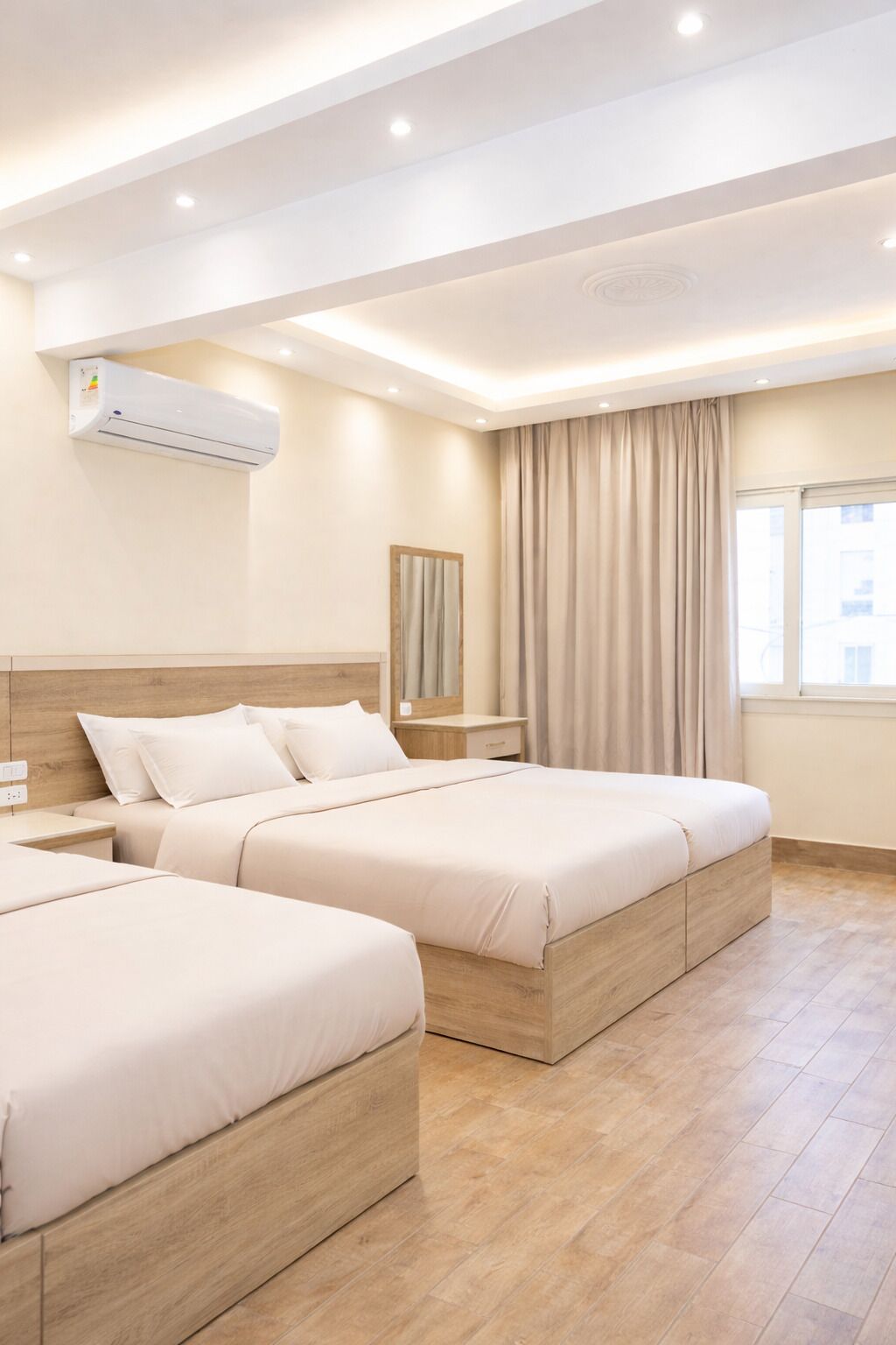 A hotel room with two beds, an air conditioner, a nightstand, a mirror, and a window with curtains.