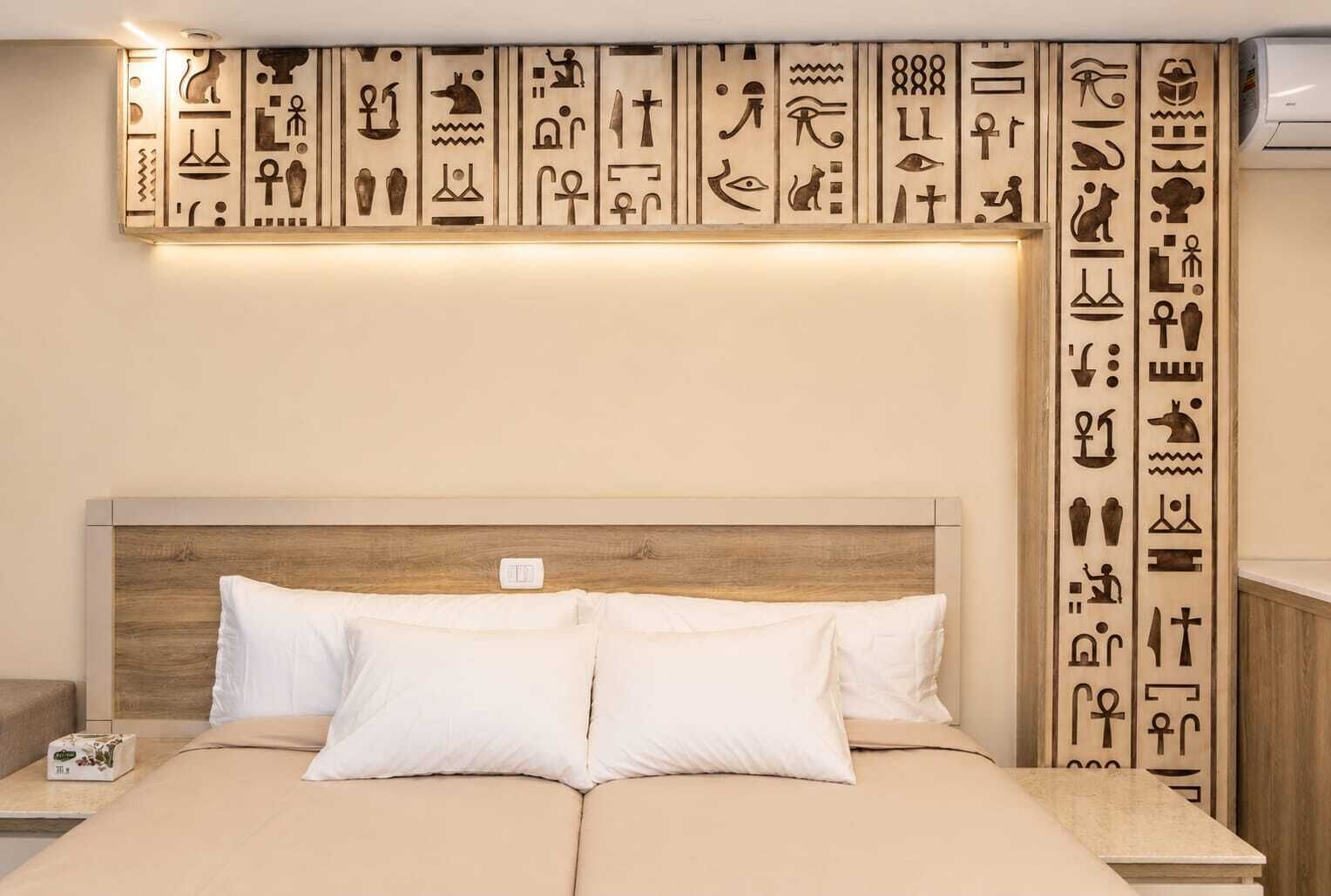 Un lit avec deux oreillers, une tête de lit en bois et un mur orné de hiéroglyphes égyptiens.