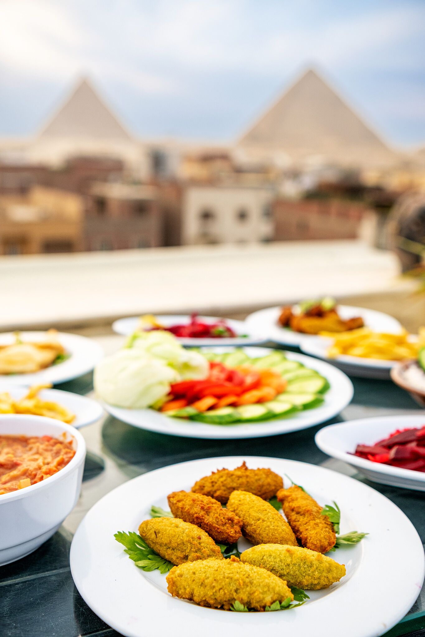 A table with various dishes, including croquettes, salad, and a bowl of sauce, set against a backdrop of pyramids.