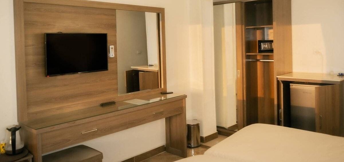 A hotel room with a wooden vanity, a flat-screen TV, a mirror, a small trash can, and a bed.