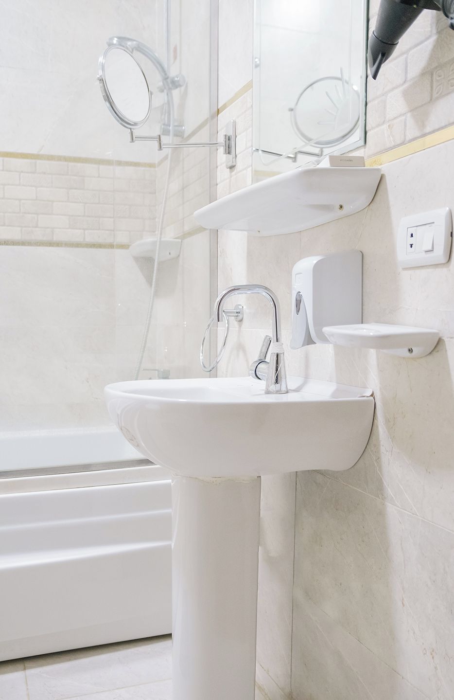A bathroom with a white pedestal sink, a handheld showerhead, and a wall-mounted soap dispenser.