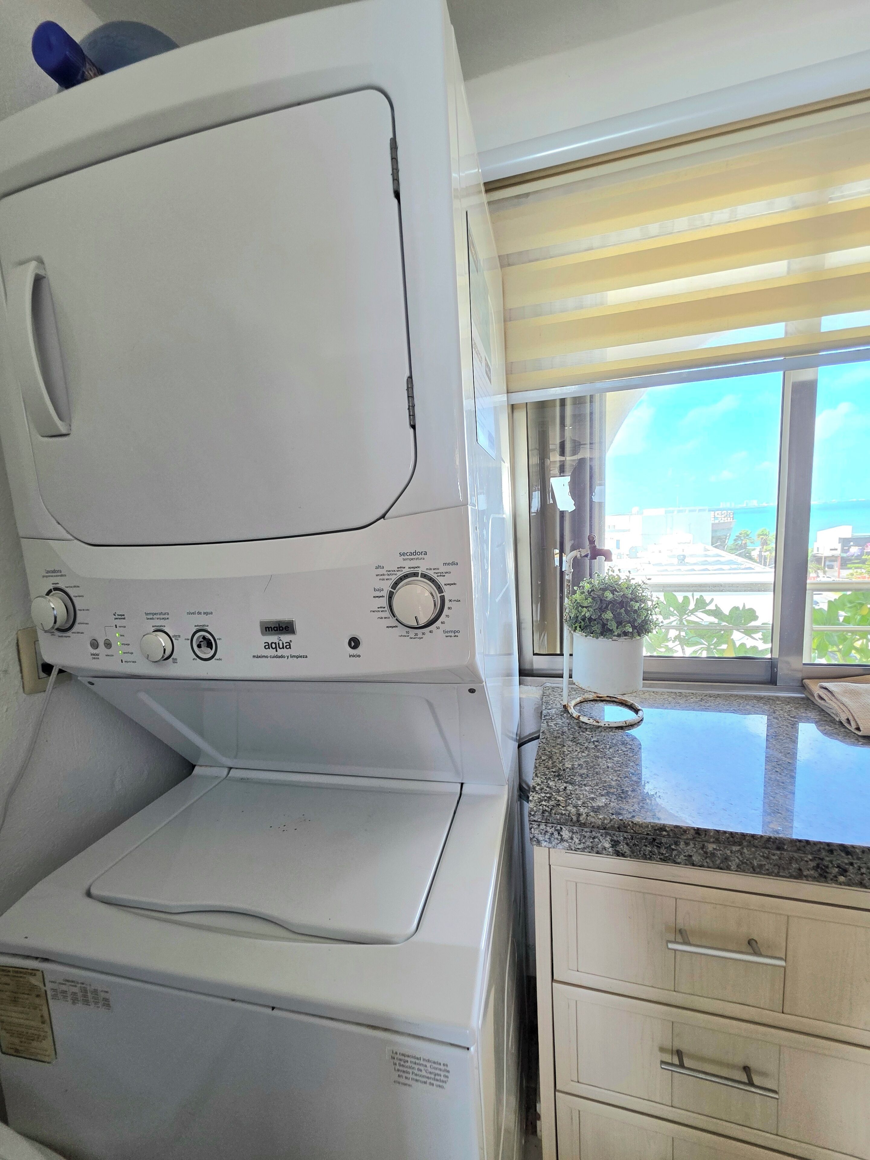 A compact, modern kitchen with a built-in washer and dryer, a granite countertop, and a view of a cityscape through large windows.