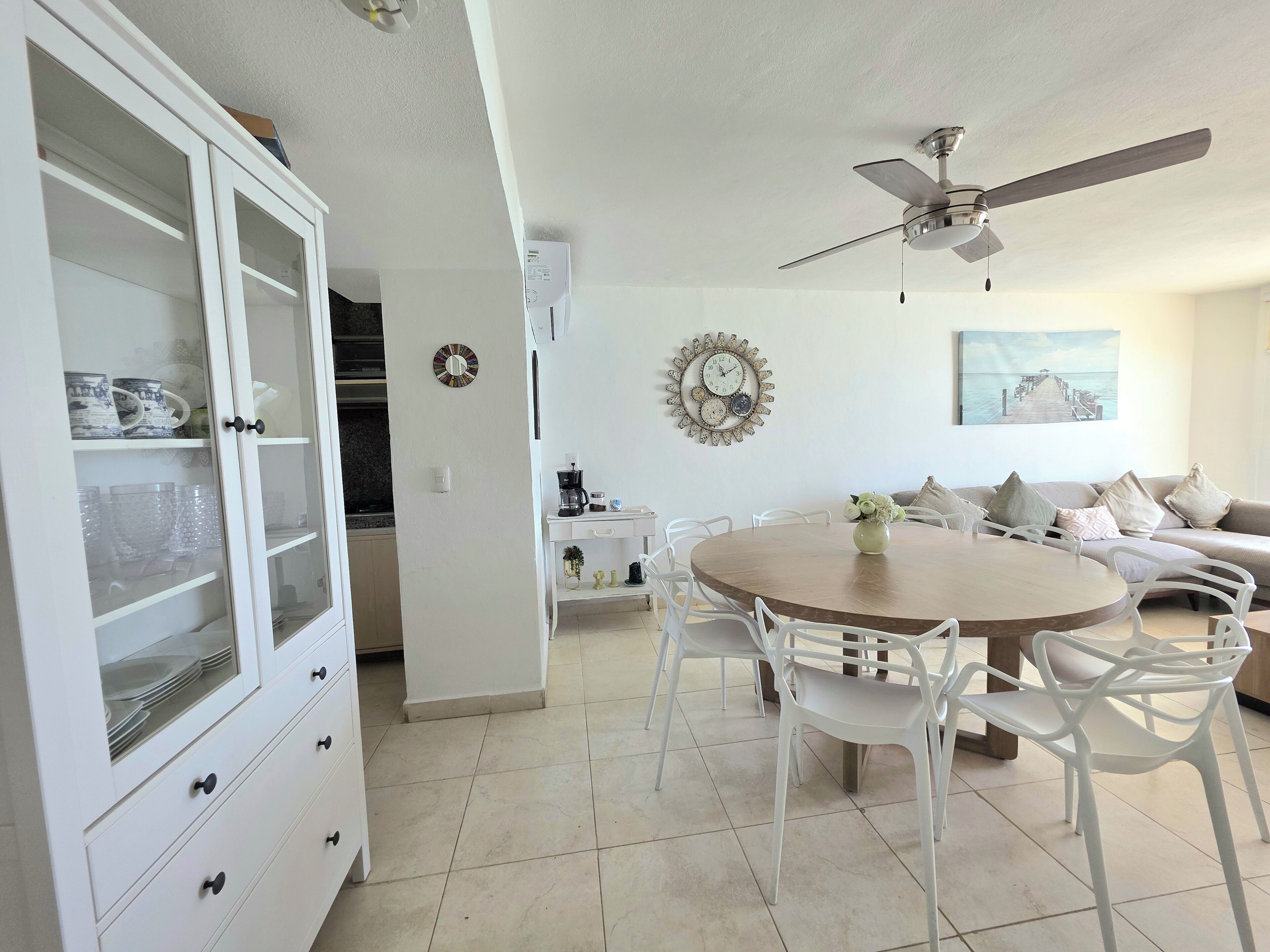 A modern kitchen with white cabinetry, a dining area with a round table and white chairs, a ceiling fan, and a wall clock.