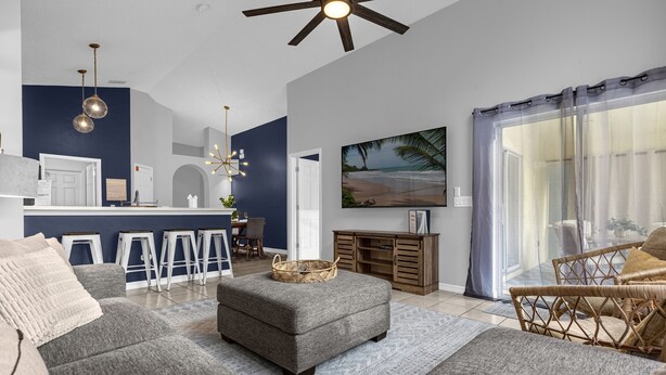 A modern living room with a grey sofa, ottoman, and wicker chair. There is a kitchen area with bar stools and a dining table. A large window with curtains is visible. - NEW*4 Bdrm, 3 Bath* Updated Detached Pool Home, 10 Minutes to Disney (Kissimmee)