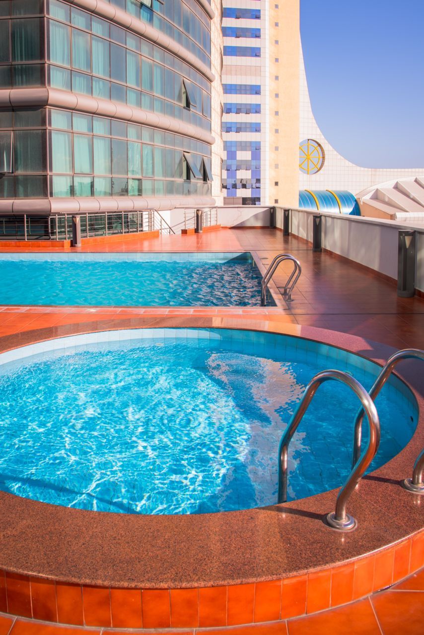 A swimming pool with a curved edge and metal railings, adjacent to a modern building with a curved facade and a logo.