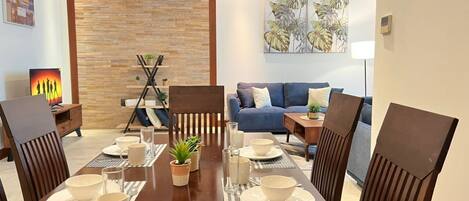 A dining area with a wooden table set for four, surrounded by chairs. There is a blue sofa and a small side table with a lamp in the background.